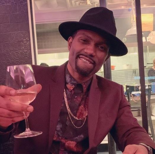 A black man drinks wine and bites his lip in a hat and suit. This man is Kaze 4 Letters