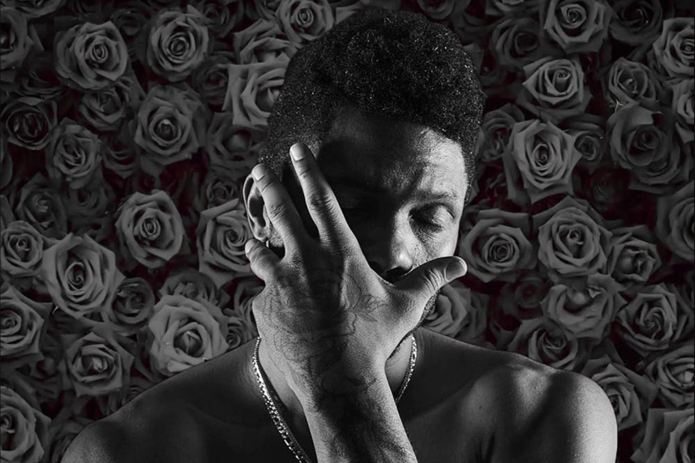 A black man covers his face with a rose tattoo on his hand and roses in the background. This man is Kaze 4 Letters